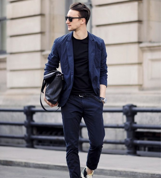 Man walking confidently in an urban setting wearing a navy blazer over a black t-shirt with matching navy trousers, black sunglasses, sneakers, and carrying a stylish black leather bag.