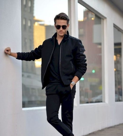 Man leaning against a white wall in an urban setting wearing an all-black outfit with a bomber jacket, slim-fit pants, sunglasses, and a wristwatch, showcasing a sleek monochrome street style.